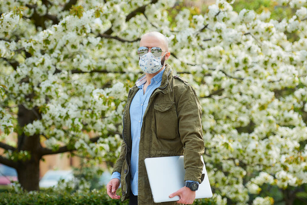 Un homme chauve avec une barbe dans un masque médical pour éviter la propagation du coronavirus marche avec un ordinateur portable dans le parc. Un mec porte un masque facial n95 et des lunettes de soleil pilotes dans le jardin entre les arbres à fleurs
. - Photo, image