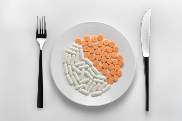 白い背景にカトラリーの横にカプセルとオレンジの錠剤が充填された白いプレートでテーブルの設定.あなたの製品のための医薬品中毒や健康的な栄養、製薬業界やイラストの概念を表すために. - 写真・画像