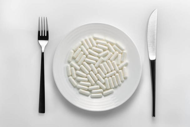 Primer plano de una mesa con un plato blanco lleno de cápsulas blancas junto a cubiertos sobre un fondo blanco. Representar un concepto de adicción farmacéutica y nutrición saludable, industria farmacéutica, ilustración para su producto
. - Foto, Imagen