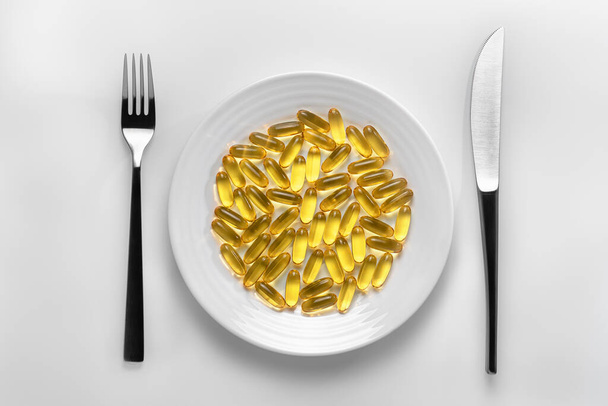 Tischdekoration mit einem weißen Teller gefüllt mit gelben Omega-3-Kapseln neben Bestecken auf weißem Hintergrund. Um ein Konzept der pharmazeutischen Abhängigkeit und gesunden Ernährung, der pharmazeutischen Industrie oder eine Illustration für Ihr Produkt darzustellen. - Foto, Bild