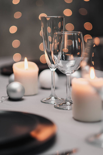 A table set in the restaurant. Serving a table. Grey artistic background. Black matte utensils, glass glasses. White candles and decor. Festive yellow lights garland in the background. - Фото, изображение