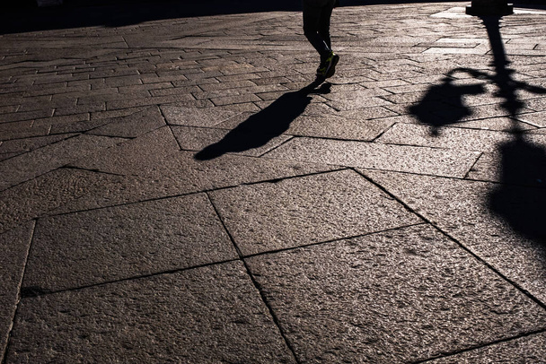 Low section of man walking on stone pavement. Abstract pattern of stone block floor with prospect in sunset light, shadow of a lonely man and a street light. - Photo, Image