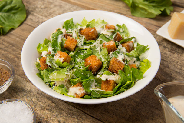 Caesar salade in een witte kom op een oude rustieke houten tafel met bladeren en een saus boot en parmezaanse kaas. Geselecteerde focus. - Foto, afbeelding