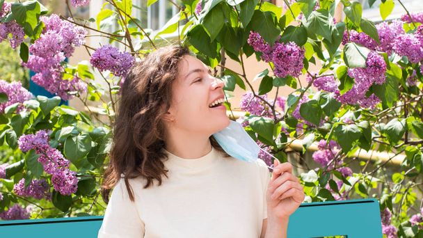Gelukkige jonge krullende vrouw die haar medisch masker afdoet en bloemen ruikt naar lila, zittend op een bank in een groene bloeiende tuin, park. Einde quarantaine, covid-19 pandemie - Foto, afbeelding