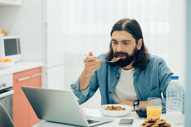 Beau jeune homme mangeant son petit déjeuner dans la cuisine et regardant un film sur un écran d'ordinateur portable
 - Photo, image