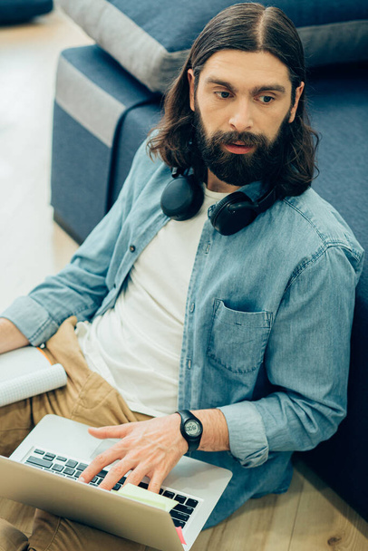 Young bearded man sitting on the floor with his back to the sofa and looking away while holding a laptop on his leg - Фото, зображення