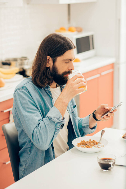 Calm handsome man at home at the kitchen table looking at the screen of his smartphone and holding a glass of juice - 写真・画像