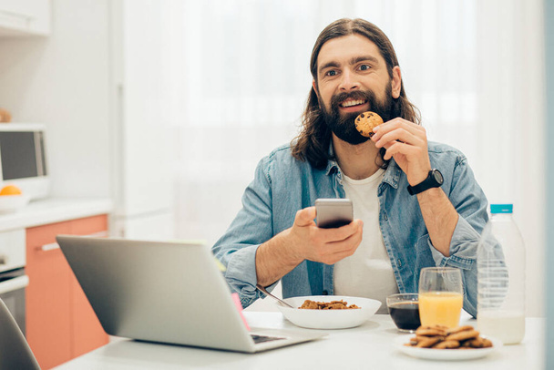 Positive young Caucasian man sitting at the kitchen table with a cookie and a smartphone in his hands - Foto, afbeelding