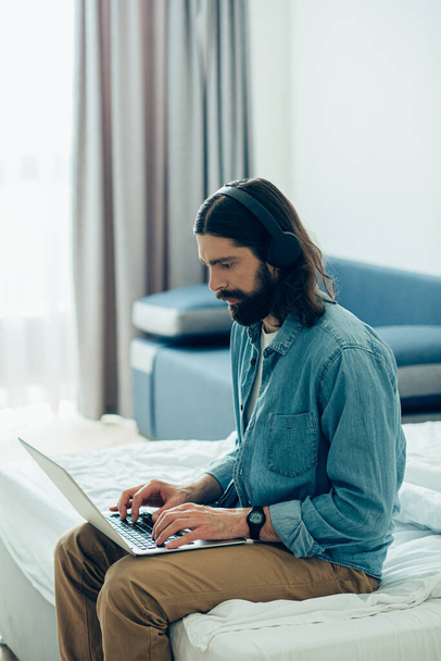 Serious bearded young man sitting on the bed and working on laptop during his self-isolation - Foto, Imagen