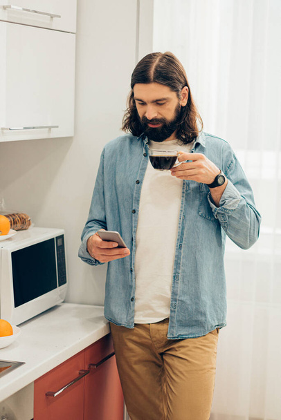 スマートフォンとコーヒーを片手に電子レンジの近くに立つカジュアルな服を着たハンサムな髭の長い髪の男 - 写真・画像