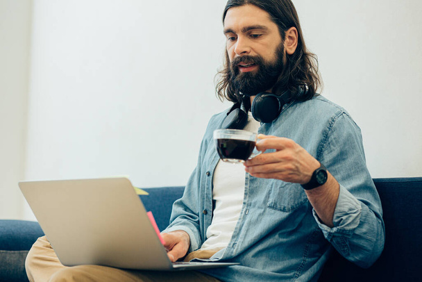 Concentrated long haired bearded man sitting in front of a laptop screen and holding a cup of coffee - Фото, изображение