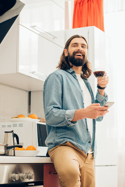 Encantado homem bonito de pé na cozinha com café e um smartphone e olhando para longe com um sorriso
 - Foto, Imagem