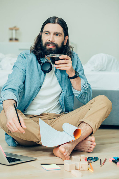 Jeune homme créatif assis pieds nus sur le sol avec des fournitures de papeterie et regardant au loin tout en buvant du café
 - Photo, image