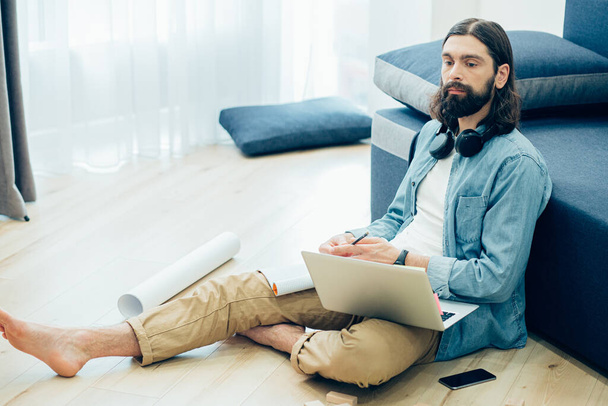 Young bearded man thoughtfully looking away while sitting on the floor in casual clothes with laptop - Φωτογραφία, εικόνα