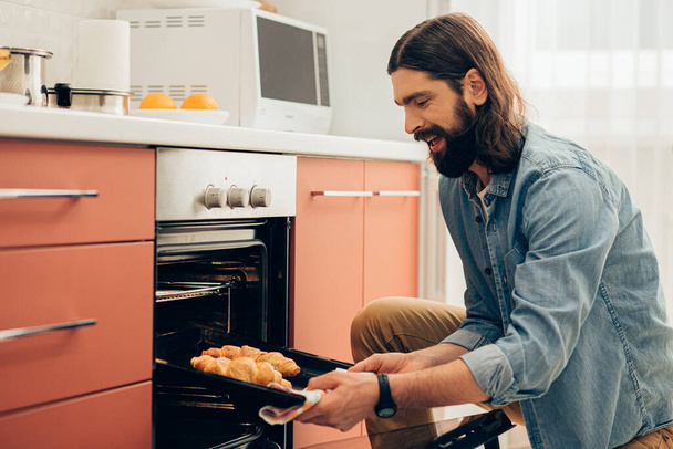 Positive young man smiling and looking impressed while taking croissants from the oven - Photo, Image
