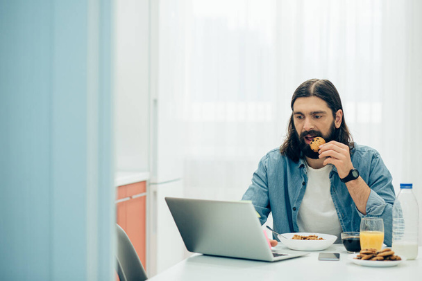 Concentrated man looking attentively at the screen and putting a cookie into his mouth - Foto, afbeelding