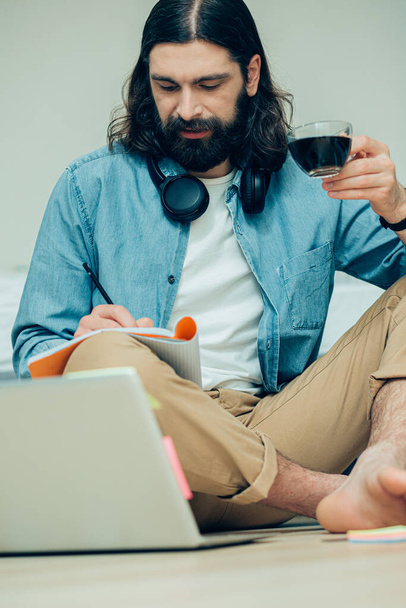 Calm young bearded man sitting in front of a laptop on the floor and making notes in a notebook - Фото, изображение