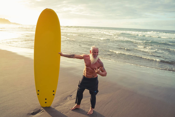 Apto para personas mayores divirtiéndose surfeando al atardecer - Entrenamiento de hombre barbudo deportivo con tabla de surf en la playa - Estilo de vida de las personas mayores sanas y concepto de deporte extremo - Imagen
 - Foto, Imagen