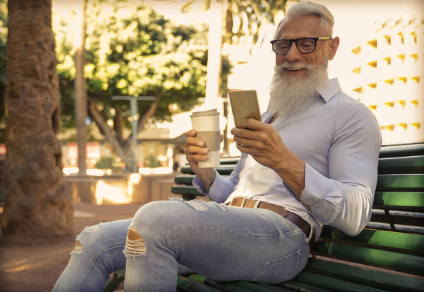 Trendy senior man using smartphone app and drinking coffee in the park. Mature fashion male having fun with new trends technology . Tech and joyful elderly lifestyle concept - Image - 写真・画像
