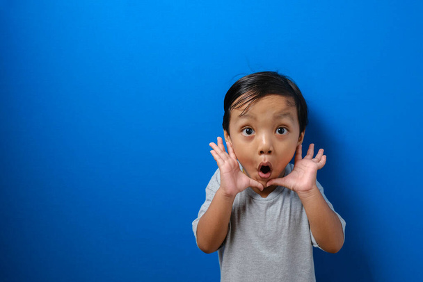 Portrait of funny young Asian boy looking at camera with big eyes covering his mouth, shocked surprised expression against blue background - Photo, Image
