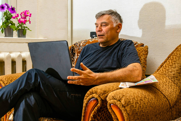 Ένας άντρας κάθεται με ένα λάπτοπ στην αγκαλιά του στο σπίτι. Πληροφοριακή έρευνα. Η επιθυμία να μάθουν και να έχουν έναν υπολογιστή στο σπίτι. Δείτε τις ειδήσεις. Μετά τη διαμονή. Προσθέτω φίλους. Προβολή δημοφιλών πόρων - Φωτογραφία, εικόνα