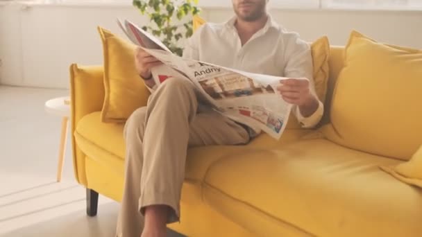 A concentrated young man is reading a newspaper while sitting on the sofa in the living room at home - Imágenes, Vídeo
