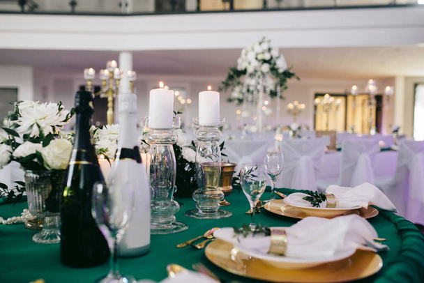 Στην αίθουσα δεξιώσεων του γάμου υπάρχουν τραπέζια με πράσινα τραπεζομάντηλα, στα τραπέζια υπάρχουν συνθέσεις από λουλούδια και χόρτα, κεριά, μαχαιροπήρουνα, στα πιάτα υπάρχουν πετσέτες, υπάρχουν δύο μπουκάλια σαμπάνιας - Φωτογραφία, εικόνα