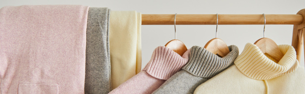 vista de cerca de suéteres suaves de punto rosa, beige y gris y pantalones colgando en estante de madera aislado en blanco, tiro panorámico
 - Foto, Imagen