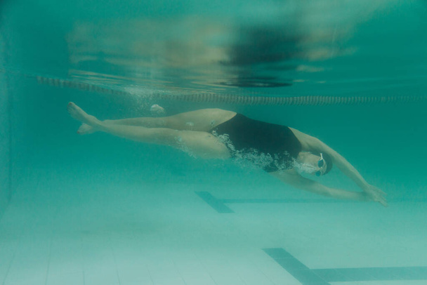 Kobieta instruktorka pływania w czarnym stroju kąpielowym, czapce i goglach pływa pod wodą - Zdjęcie, obraz