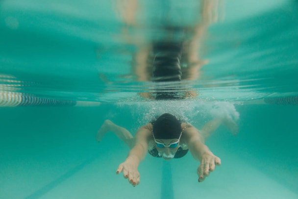 Female swimming instructor in a black swim suit, a cap and goggles is swimming under water - Fotoğraf, Görsel