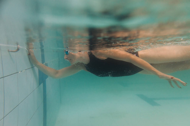 Schwimmlehrerin in schwarzem Badeanzug, Mütze und Schutzbrille schwimmt unter Wasser - Foto, Bild