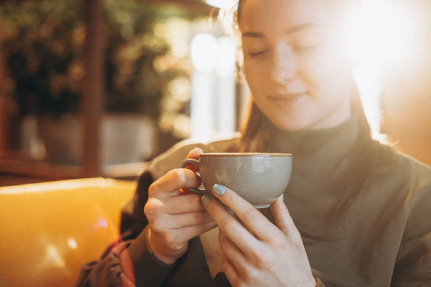 young pretty girl with a big mug of sweet coffee sits on a chair in her favorite cafe and drinks her daily morning coffee and talks with a girlfriend - 写真・画像