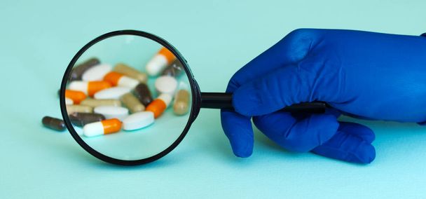 kéz a kék orvosi kesztyű tartja nagyító néz fehér kerek tabletta, közelről, szelektív fókusz, elmosódott semleges háttér - Fotó, kép