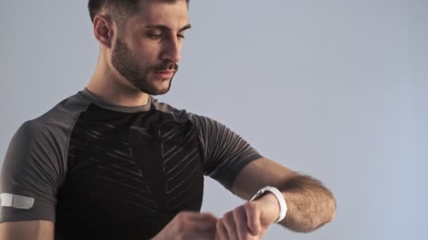 A calm young man in sportswear is using his fitness tracker while doing working out exercises isolated over the grey background in studio - Filmati, video