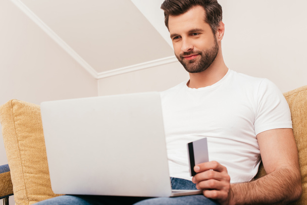 Επιλεκτική εστίαση του ανθρώπου χρησιμοποιώντας φορητό υπολογιστή και κρατώντας πιστωτική κάρτα στο σπίτι  - Φωτογραφία, εικόνα