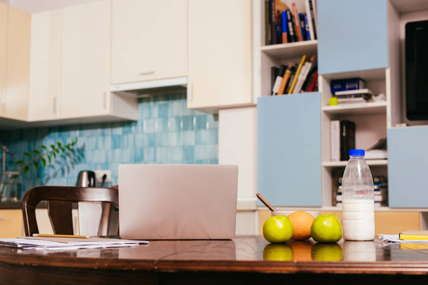 Χαρτιά με φορητό υπολογιστή κοντά σε φρούτα και πρωινό στο τραπέζι στην κουζίνα  - Φωτογραφία, εικόνα