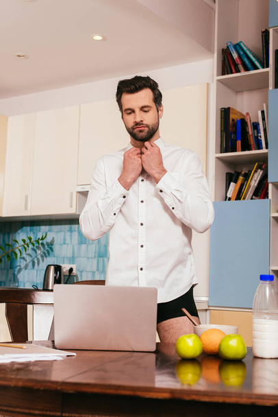 Вибірковий фокус красивого чоловіка в трусиках надягаючи сорочку біля ноутбука і сніданок на кухонний стіл
  - Фото, зображення