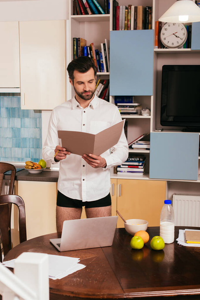 Selektiver Fokus eines gutaussehenden Mannes in Hemd und Höschen, der mit Papieren in der Nähe von Laptop und Frühstück auf dem Tisch arbeitet  - Foto, Bild
