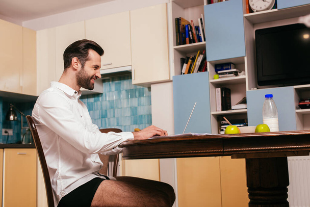 Side view of smiling man in shirt and panties using laptop near breakfast on table in kitchen  - Fotoğraf, Görsel