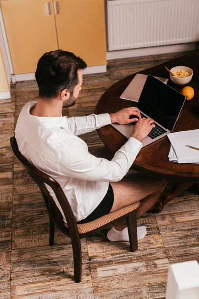 Overhead view of man in panties and shirt using laptop near papers and cereals on table  - Fotografie, Obrázek