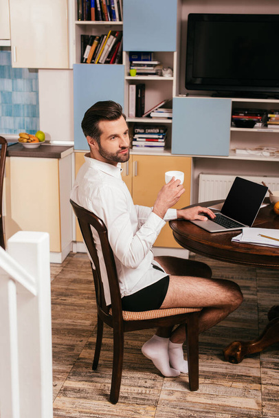 Válogatott fókusz jóképű férfi ing és bugyi segítségével laptop és tartó csésze a nappaliban - Fotó, kép
