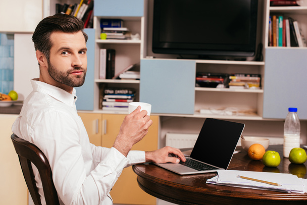 Vista lateral del hombre guapo sosteniendo taza de café cerca de la computadora portátil y papeles en la mesa
  - Foto, imagen