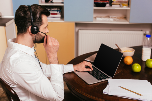 Oldalnézet férfi headset dolgozik laptop közelében reggeli az asztalon  - Fotó, kép