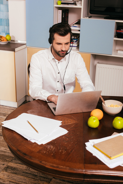 Komea mies kuulokkeissa kannettavan tietokoneen lähellä aamiaista pöydällä
  - Valokuva, kuva