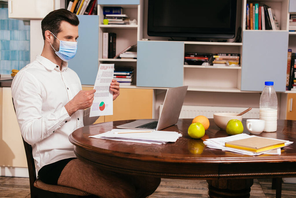Freelancer en máscara médica, camisa y bragas que sostiene el documento mientras tiene video chat en el portátil cerca del desayuno en la mesa
  - Foto, Imagen
