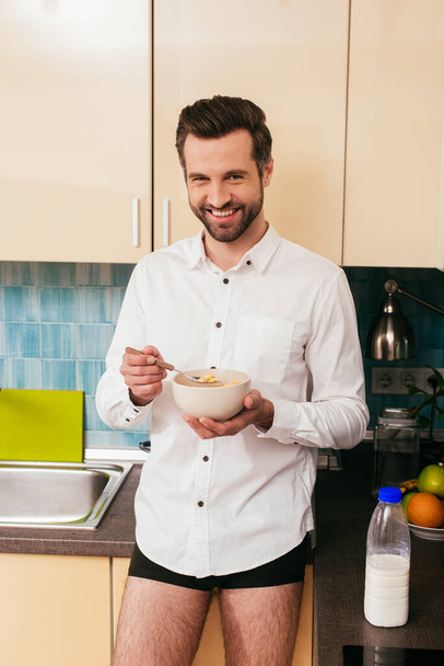 Handsome man in shirt and panties smiling at camera while eating cereals in kitchen  - Fotó, kép
