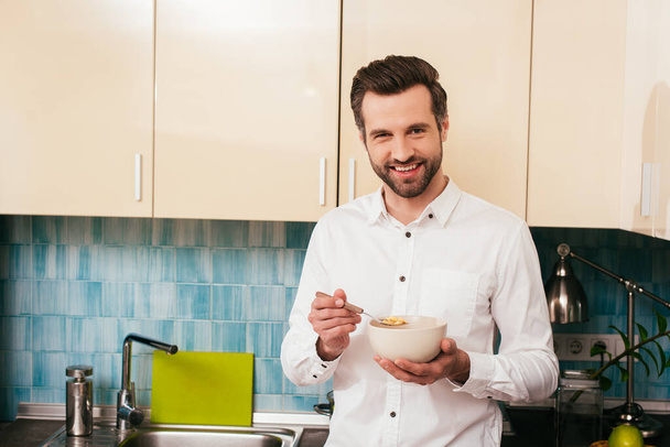 Jóképű férfi mosolyog a kamerába, miközben gabonát eszik a konyhában.  - Fotó, kép