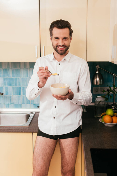 Positive man in shirt and panties holding bowl of cereals in kitchen  - Fotografie, Obrázek