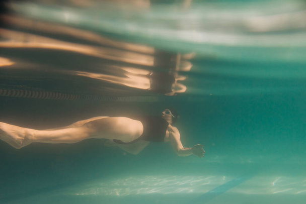 Женщина-инструктор по плаванию, находящаяся под водой, носит очки, черный купальник и кепку
 - Фото, изображение