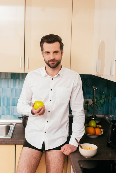 Handsome man in panties and shirt holding apple near cereals on kitchen worktop  - Photo, Image